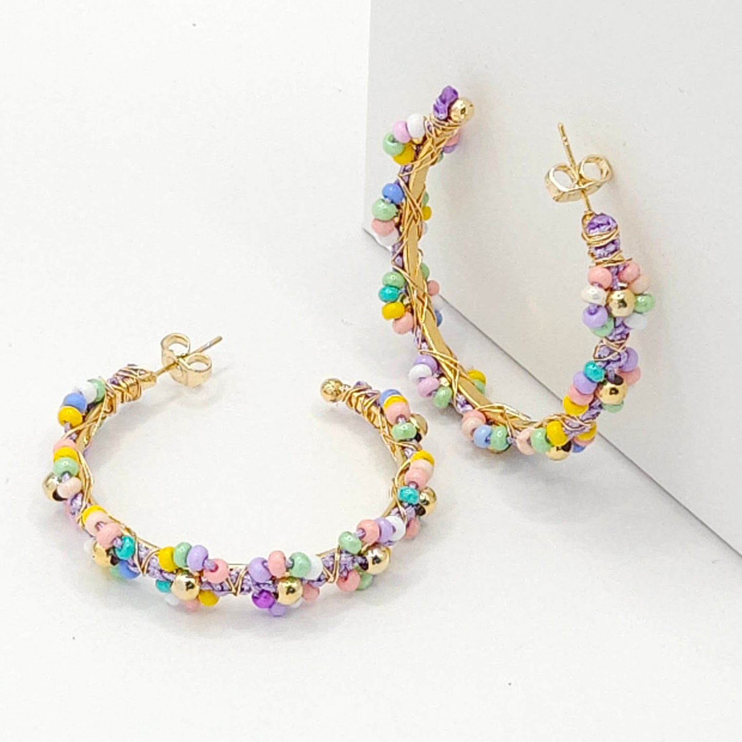 Golden hoops with soft pastel Czech beads, one lying on a white base, the other standing against a white wall
