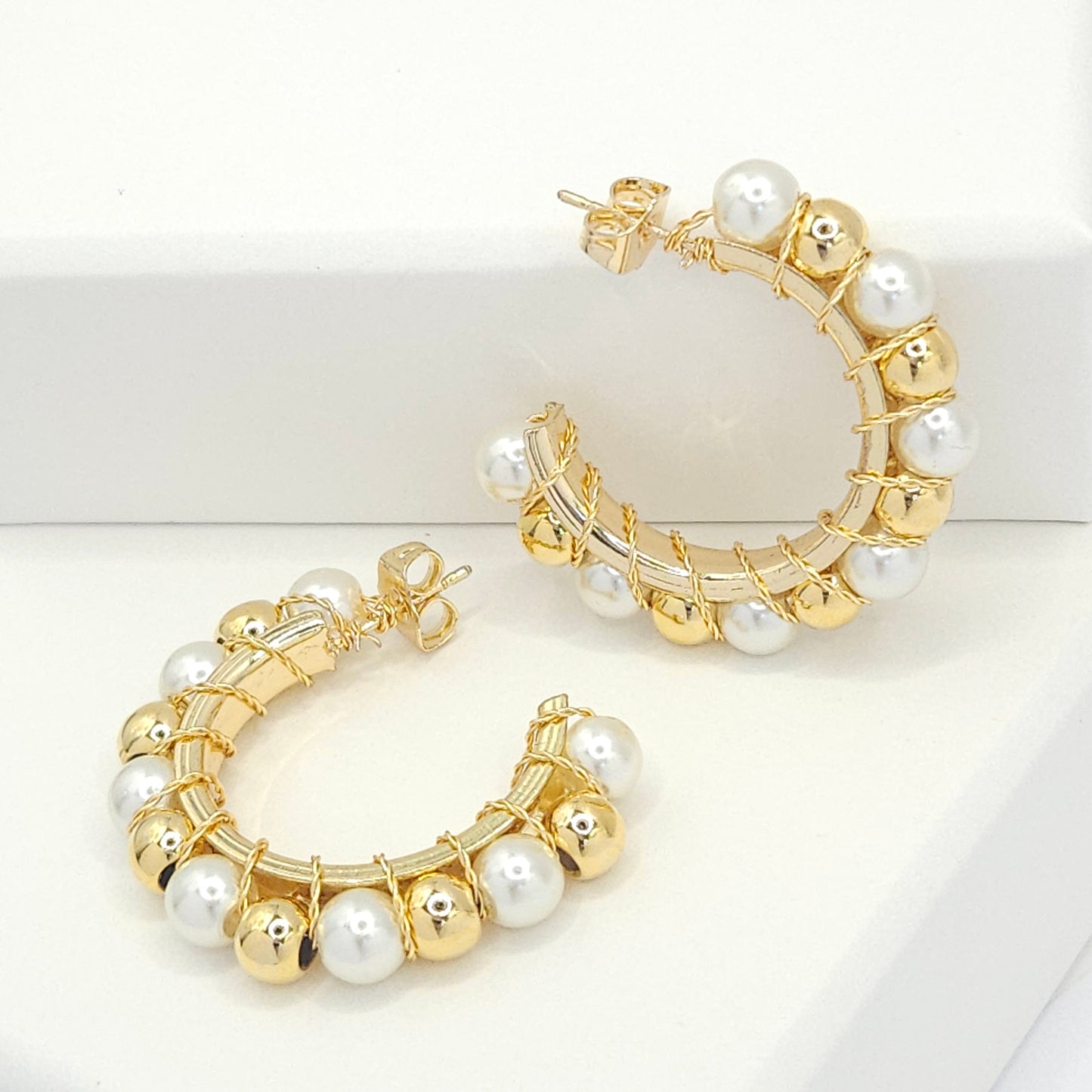 Golden hoops with pearls and golden spheres, one lying on a white base and the other standing against a white box
