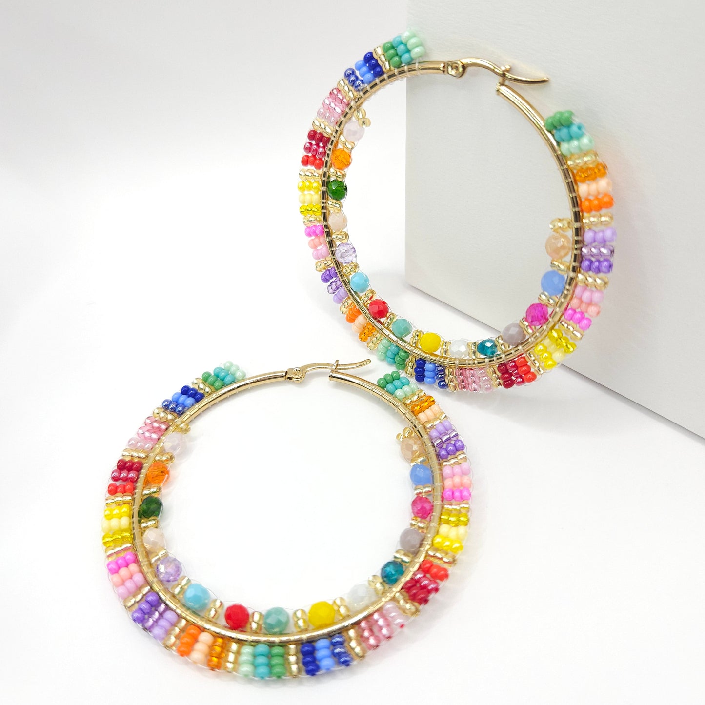 Golden hoops with colorful Czech and faceted beads, one lying on a white base, the other standing against a white wall