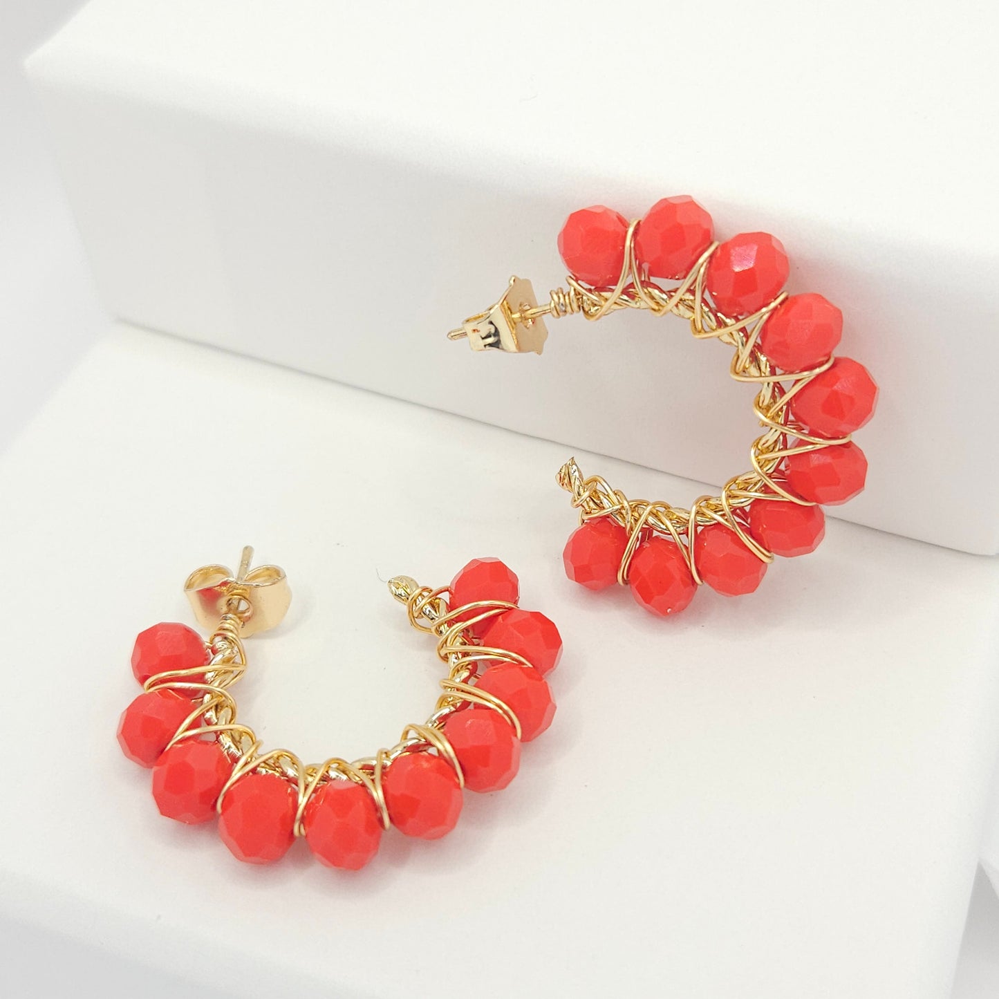 Golden hoops with delicate red faceted rondelle glass beads, one lying on a white base and the other standing against a white box