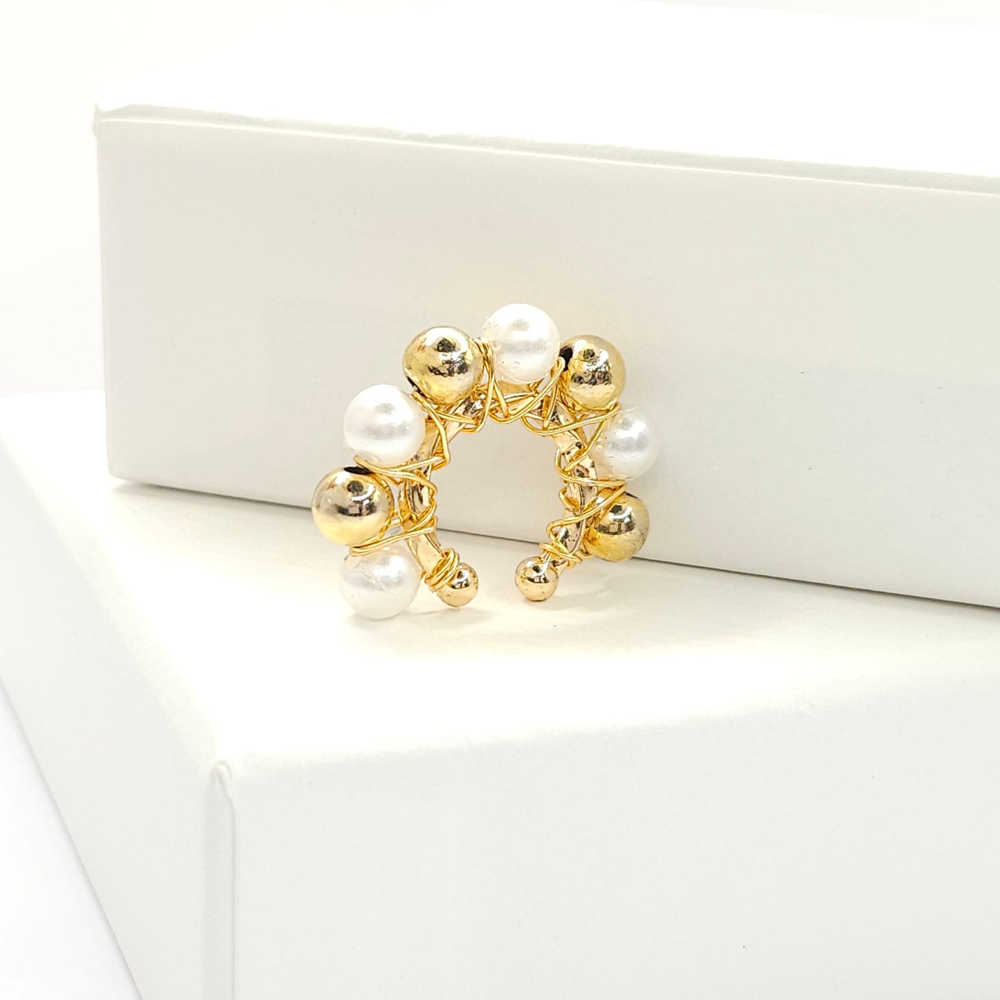 Golden ear cuff adorned with golden spheres and white pearls standing on a white base