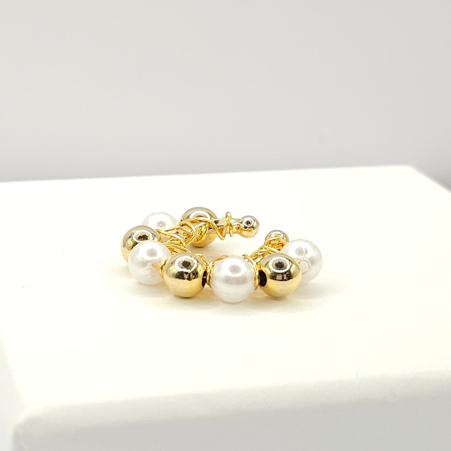 Golden ear cuff adorned with golden spheres and white pearls lying on a white base