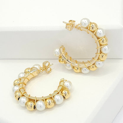 Golden hoops with pearls and golden spheres, one lying on a white base and the other standing against a white box
