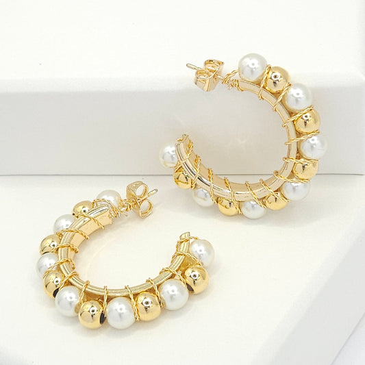 Golden hoops with pearls and golden spheres, one lying on a white base and the other standing against a white box