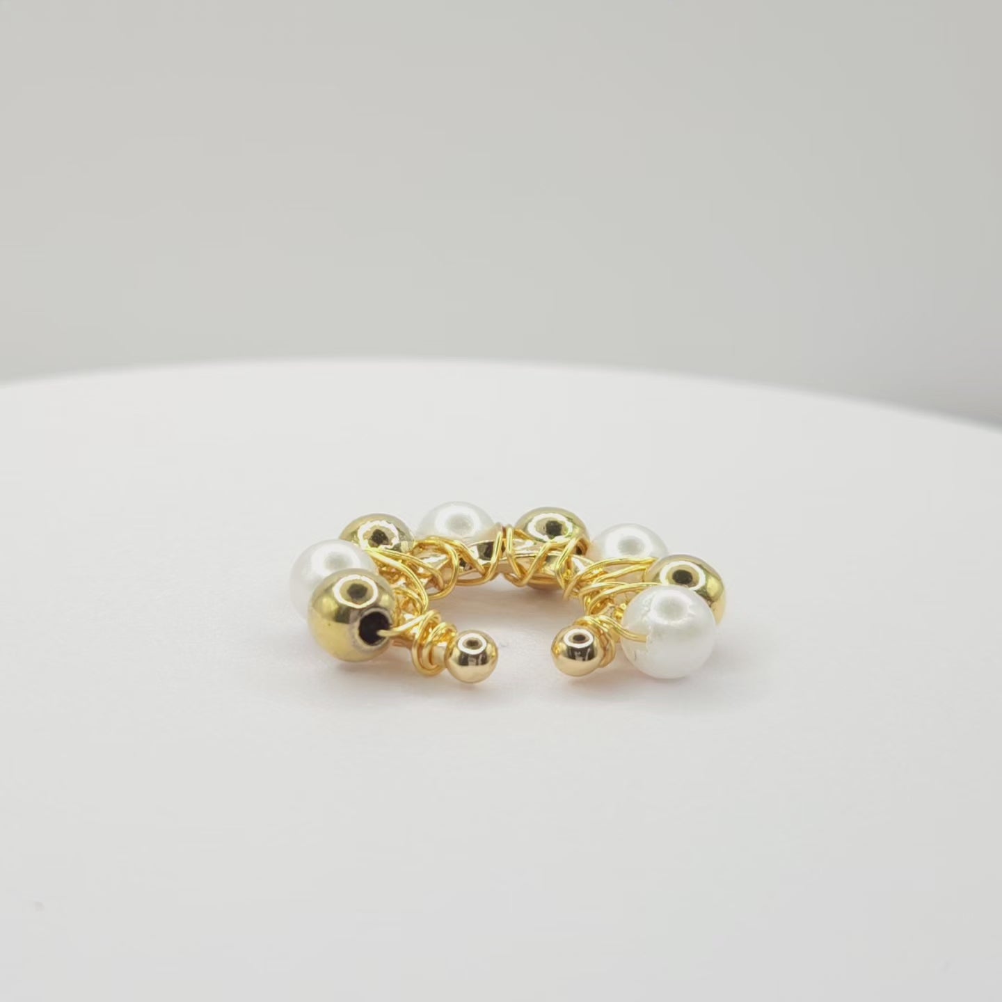 Video of a golden ear cuff adorned with golden spheres and white pearls standing on a white base, rotating counterclockwise