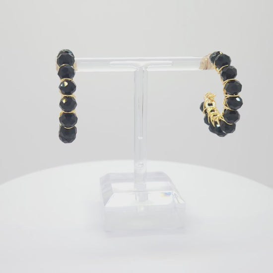 Video of golden hoops with delicate black faceted rondelle glass beads, both standing on a white base rotating counterclockwise