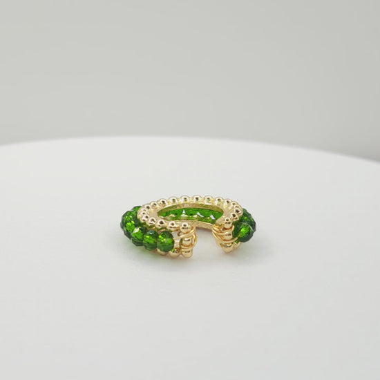 Video of a golden ear cuff featuring an array of green faceted rondelle glass beads lying on a white base, rotating counterclockwise