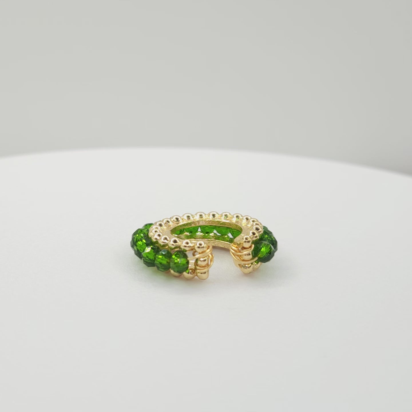 Video of a golden ear cuff featuring an array of green faceted rondelle glass beads lying on a white base, rotating counterclockwise
