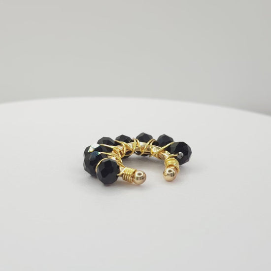 Video of a golden ear cuff adorned with a weave of black faceted rondelle glass beads on a white base, rotating counterclockwise