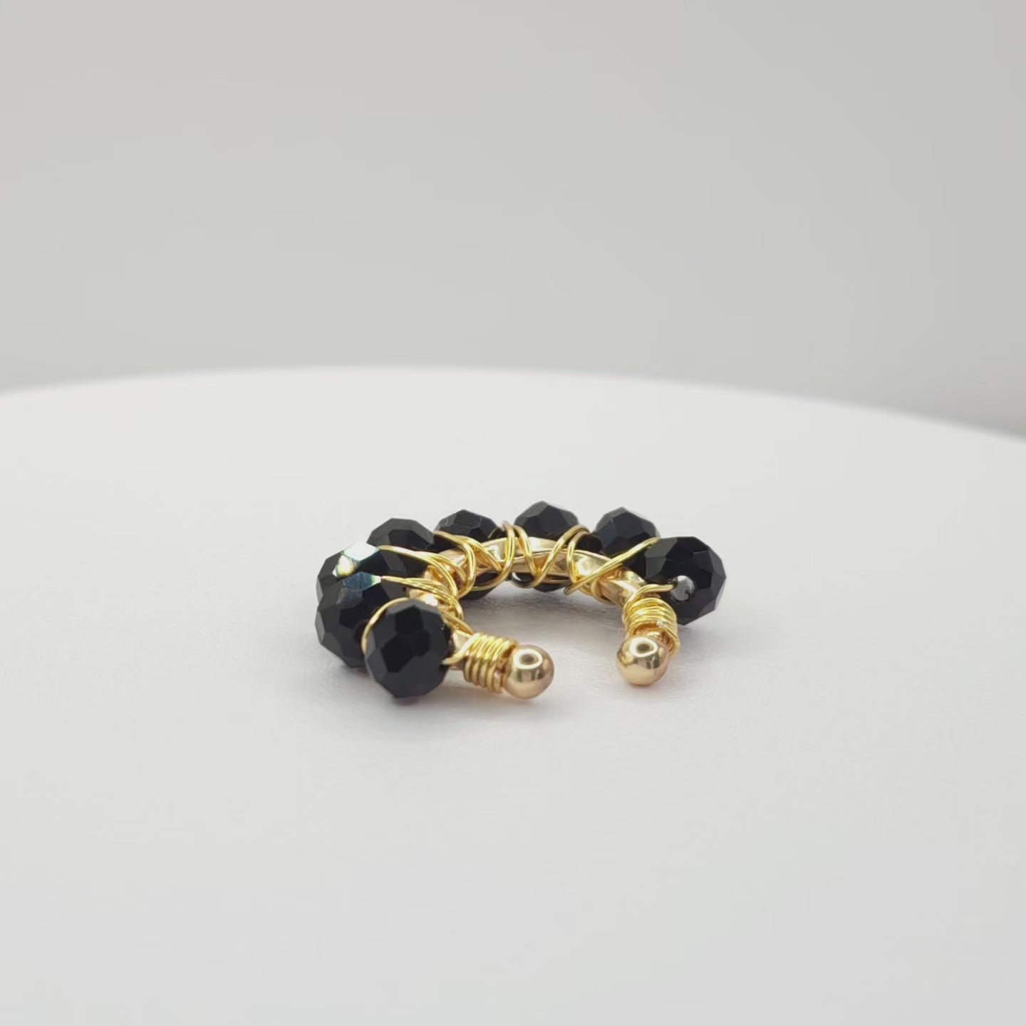 Video of a golden ear cuff adorned with a weave of black faceted rondelle glass beads on a white base, rotating counterclockwise