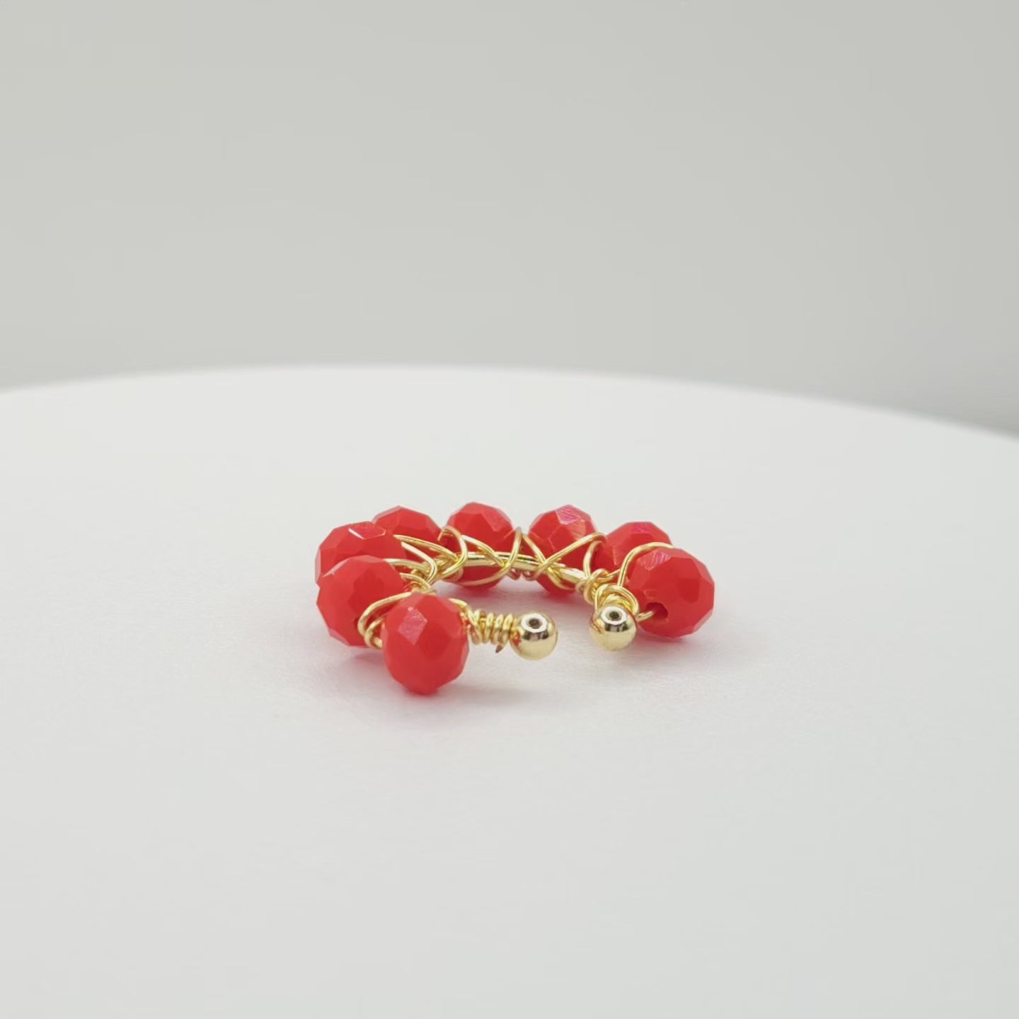 Video of a golden ear cuff adorned with a weave of red faceted rondelle glass beads on a white base, rotating counterclockwise